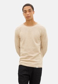 Hellbeige, langärmliger Strickpullover mit Rundhalsausschnitt und Raglanärmeln, kombiniert mit schwarzen Hosen. Weiche Textur mit einem dezenten Muster.