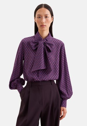 Femme portant un chemisier violet à motifs avec un grand nœud au cou et un pantalon foncé taille haute, debout avec une main dans la poche.