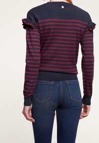 Merinovihreä ja viininpunaraitainen neulepaita, jossa on röyhelöhiha, ribattužhelma ja napitus takana. Yhdistetty tummansiniset farkut.