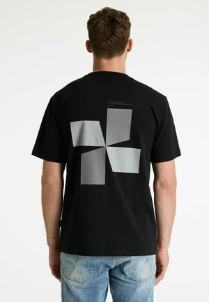 Férfi fekete pólót visel, hátul szürke, absztrakt, négypaneles grafikával, világoskék farmerrel párosítva.