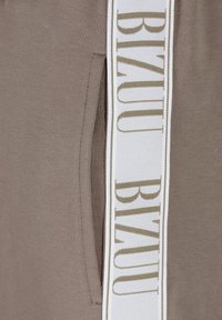 Bruine jogger van stof met een zijzak, voorzien van een witte taillebandlabel met "BIZUU" in gouden letters. Zachte textuur, minimalistisch ontwerp.