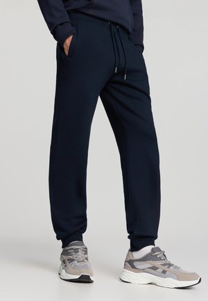 Pantaloni sportivi blu navy con vita elastica e cordino. Presentano tasche laterali e polsini a coste. Abbinati a scarpe da ginnastica grigio chiaro.
