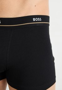 Boxer-briefs noirs en coton avec une large ceinture élastique arborant un logo "BOSS" blanc et une ligne dorée en accent. Texture lisse.