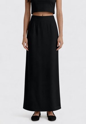 Donna che indossa una gonna lunga nera a vita alta abbinata a un top nero corto e scarpe nere basse, in piedi davanti a uno sfondo semplice.