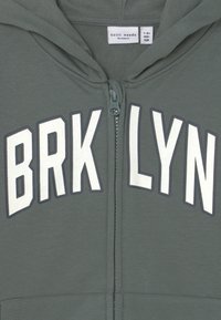 Sudadera verde con cremallera, que presenta "BRKLYN" en letras grandes blancas, con una textura suave y capucha ajustable con cordones. Etiqueta visible en el interior.