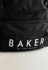 Schwarzer Stoffrucksack mit einem auffälligen weißen "BAKER"-Logo und einem kleineren Text "by TED BAKER". Enthält eine Reißverschlusstasche auf der Vorderseite.