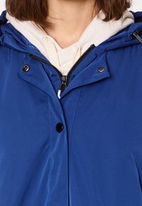 Blå huvjacka med en slät, vattentät finish, med en dragkedja och tryckknappar vid halsen, lager över en lätt hoodie.