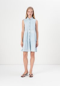 Ανοιχτό μπλε τζιν φόρεμα χωρίς μανίκια με γιακά, μπροστινές κουμπώνες και μαζεμένη μέση. Η φουσκωτή φούστα τελειώνει πάνω από το γόνατο. Φοριέται με μαύρες σαγιονάρες.