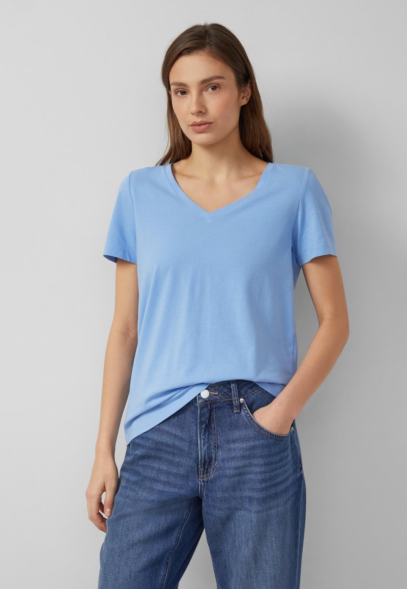 Junge Frau trägt ein hellblaues T-Shirt mit V-Ausschnitt und blaue Jeans, steht mit einer Hand in der Tasche vor einem einfarbigen Hintergrund.