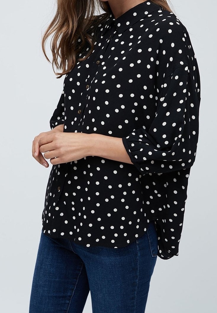 Femme portant une chemise noire à pois blancs boutonnée et un jean bleu, les mains légèrement jointes devant, sur un fond uni.