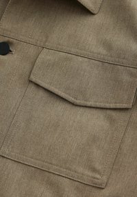 Primă de țesătură a unei jachete khaki, cu un buzunar pătrat pentru piept, cusături vizibile și o textură netedă. Accent de nasture negru vizibil.