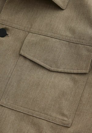 Primo piano del tessuto di una giacca khaki che presenta una tasca quadrata sul petto, cuciture visibili e una consistenza liscia. Accento del bottone nero ben in evidenza.