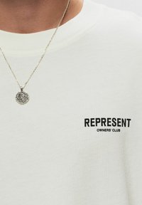 Camiseta de algodón color crema con el texto "REPRESENT OWNERS' CLUB" en negro. La tela tiene una textura suave y lisa. Collares de plata visibles.