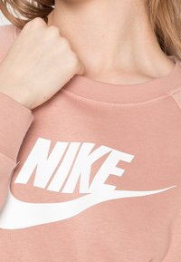 Ljusrosa sweatshirt med rund halsringning och ribbade ärmslut, med en framträdande vit Nike-logotyp och swoosh-design på framsidan.