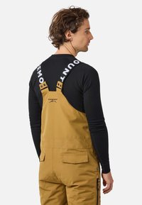 Mężczyzna w żółtych ogrodniczkach na ramiączkach, czarny długi rękaw pod spodem, stoi bokiem na białym tle.