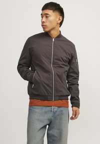 Dark gray bomber jacket with a zip front, ribbed cuffs, side pockets, and a smooth texture, worn over an orange shirt and light blue jeans.