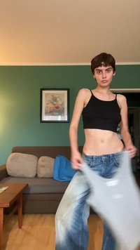 Top cropped preto com alças finas, combinado com jeans de perna larga desfiados em azul claro. Sofá em tons neutros ao fundo.