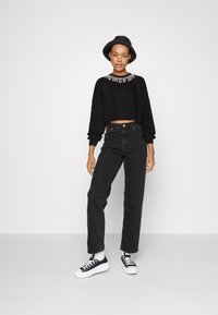 Sudadera nera corta con scollatura decorata, abbinata a jeans neri a vita alta e sneakers nere con suola bianca. Indossa un cappello nero.