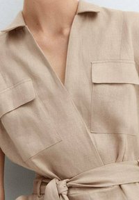 Robe portefeuille en lin beige avec des manches courtes, deux poches poitrine et une ceinture à nouer à la taille, présentant une texture lisse et un col classique.