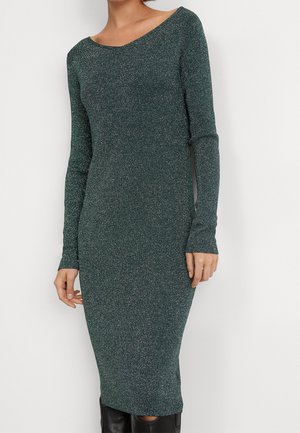 Vrouw draagt een aansluitende, lange mouwen, donkergroene glitter gebreide jurk met een brede halslijn en zwarte kniehoge laarzen.