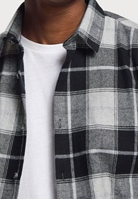 Černá a šedá kostkovaná flanelová košile s knoflíkovým zapínáním, rozšířeným límcem a kontrastním bílým tričkem pod ní; měkká textura, lehká látka.