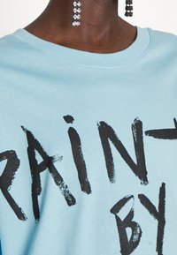 Himmelblaues Baumwoll-Sweatshirt mit großen schwarzen Pinselstrich-Text "PAINT BY." Mit kurzen Ärmeln und Rundhalsausschnitt.