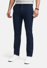 Chino navy închis, cu o croială slim, realizat dintr-un material texturat, cu buzunare frontale și accente laterale minime. Asortat cu adidași albi.