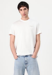 Weißes Baumwoll-T-Shirt mit Rundhalsausschnitt und kurzen Ärmeln, versehen mit einem kleinen "TOMMY JEANS"-Logo auf der linken Brust. Getragen mit hellblauen Jeans.