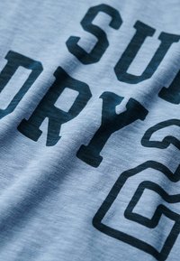 Lichtblauw katoenen t-shirt met donkere bedrukt tekst met een lichte textuur. Het ontwerp bevat vetgedrukte letters en cijfers in een casual stijl.