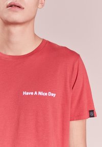 Young person wearing a red t-shirt with white text "Have A Nice Day" on the chest and a small black tag on the sleeve.