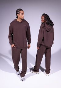Sudadera con capucha marrón y pantalones a juego, de tejido suave y corte relajado, con un pequeño logo en el pecho y detalles de costura sutiles.