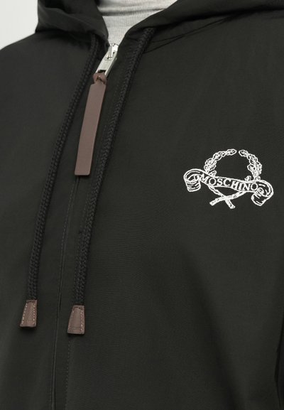 Črna zip-up kapuca z sprednjim žepom, vrvico za nastavitev in belo vezeno logotipno zasnovo. Gladek material z mat površino.