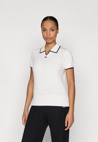 Λευκό πόλο μπλουζάκι με μαύρη περιφέρεια στο γιακά και ένα μικρό λογότυπο στο στήθος. Λείος υφασμάτινος τύπος και κοντά μανίκια. Συνδυάζεται με μαύρο πλεκτό παντελόνι.