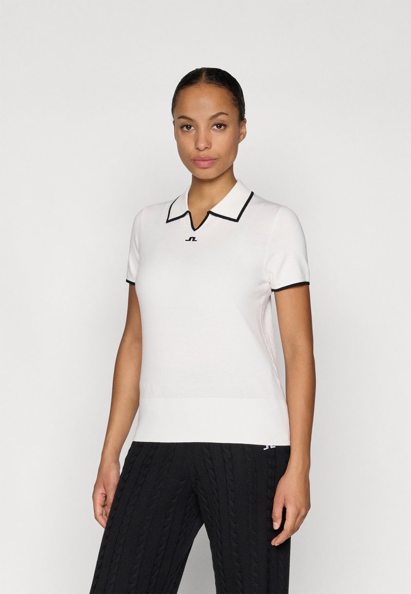 Λευκό πόλο μπλουζάκι με μαύρη περιφέρεια στο γιακά και ένα μικρό λογότυπο στο στήθος. Λείος υφασμάτινος τύπος και κοντά μανίκια. Συνδυάζεται με μαύρο πλεκτό παντελόνι.