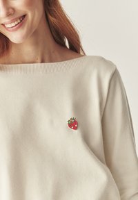 Pull crème avec un décolleté large, présentant un petit design de fraise brodé sur le devant, fabriqué en matière tricotée douce.