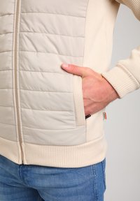 Svetlo bež zip-up jakna s quiltanimi paneli, rebrastimi manšetami in robom, stranskimi žepi in gladko teksturo blaga.