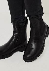 Mustat nahkaiset nilkkurit, joissa on krokotiilinaurista textuuri, joustavat sivupaneelit ja kestävä kumipohja. Suunnittelu on tyylikäs ja moderni.