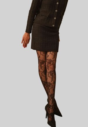 Frau in einem schwarzen strukturierten Hosenanzug, floral gemusterten schwarzen Spitzenstrumpfhosen, mit hohen Schuhen und einem leicht nach hinten ausgestreckten Arm.