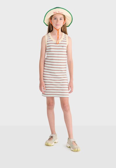 Jeune fille portant une robe blanche sans manches à rayures rouges, un chapeau de soleil beige avec une sangle orange sous le menton et des baskets beiges.