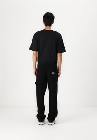Maglietta nera a maniche corte e pantaloni cargo neri. I pantaloni sono dotati di più tasche. Il modello indossa sneakers bianche e verdi. Sfondo bianco.