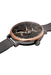 Reloj con correa de malla negra que presenta una esfera transparente con acentos dorados, mostrando intrincados engranajes y una caja negra suave y redondeada.