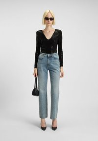 Vrouw in een zwart pailletten top, lichtblauwe hoge taille jeans met rhinestones, zwarte hakken, zonnebril en een zwarte handtas.