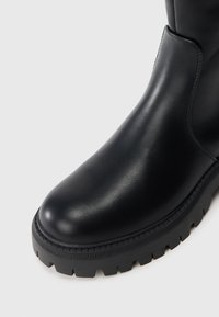 Botină neagră din piele la gleznă, cu vârf rotund, talpă din cauciuc texturat și detalii de cusătură pe margine. Finish neted și design robust.