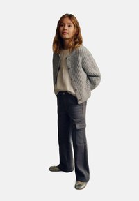 Grauer Strickcardigan mit Knopfleiste, darunter ein weißes, strukturiertes Shirt und dunkle Cargo-Hosen. Das Outfit wird mit silbernen Schuhen abgerundet.