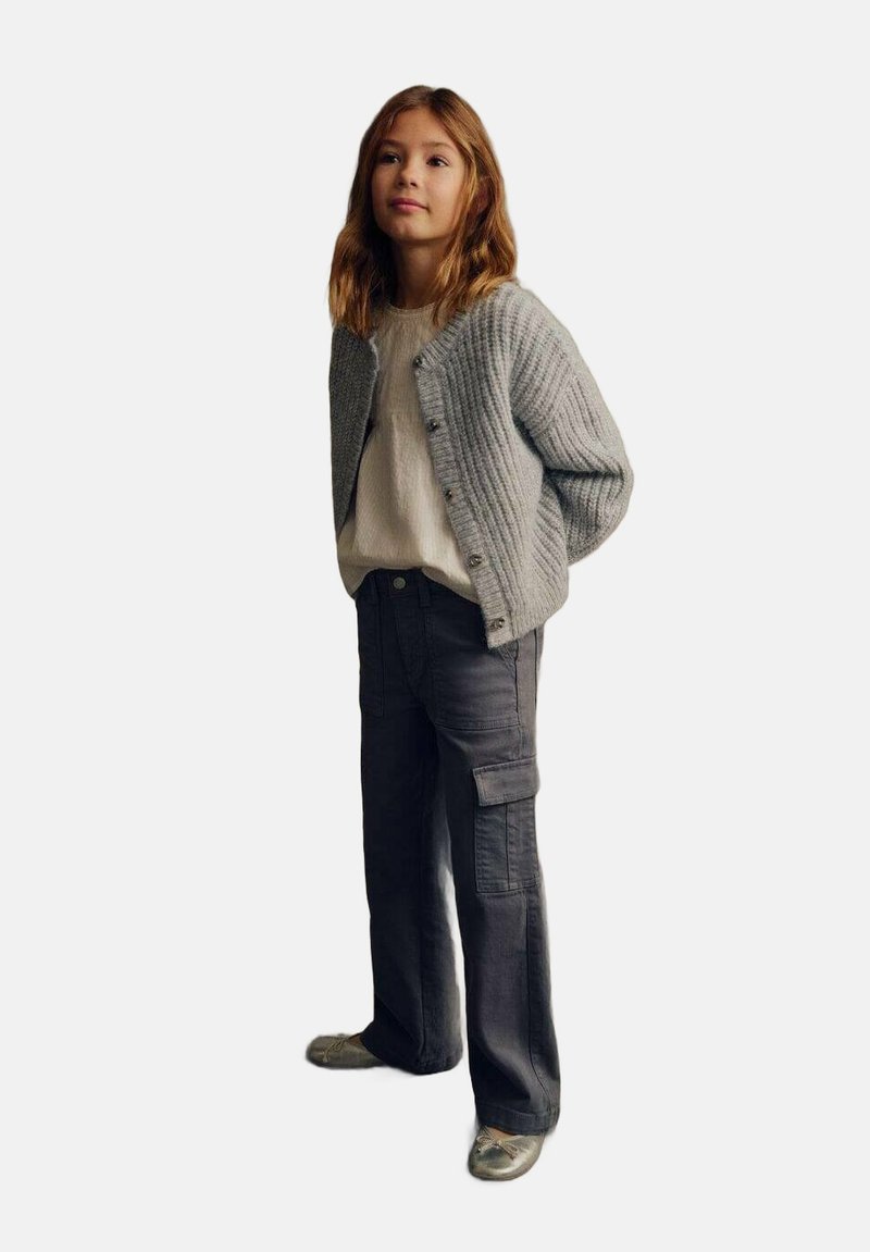 Grauer Strickcardigan mit Knopfleiste, darunter ein weißes, strukturiertes Shirt und dunkle Cargo-Hosen. Das Outfit wird mit silbernen Schuhen abgerundet.