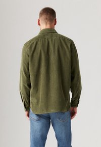 Camicia in velluto a coste verde con colletto button-down e maniche lunghe, caratterizzata da un orlo dritto e una sottile texture. Indossata con jeans in denim.