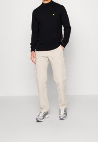 Pull à col roulé en tricot noir avec un logo jaune, associé à un pantalon cargo beige et des chaussures de sport grises avec des accents argentés.