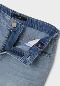 Jeans de mezclilla azul claro con una cremallera abierta y un botón, que muestran una cintura elástica y una pequeña etiqueta de marca negra en el interior.