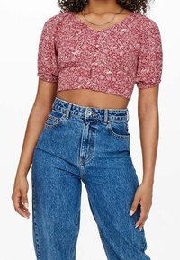 Rotes Blumen-Crop-Top mit kurzen Puffärmel, V-Ausschnitt und Knopfverschluss, kombiniert mit hochgeschnittenen blauen Jeans.