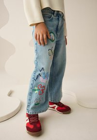 Jasnoniebieskie dżinsy z kolorowymi haftowanymi wzorami motyli i kwiatów na nogawkach, noszone z czerwono-białymi tenisówkami.